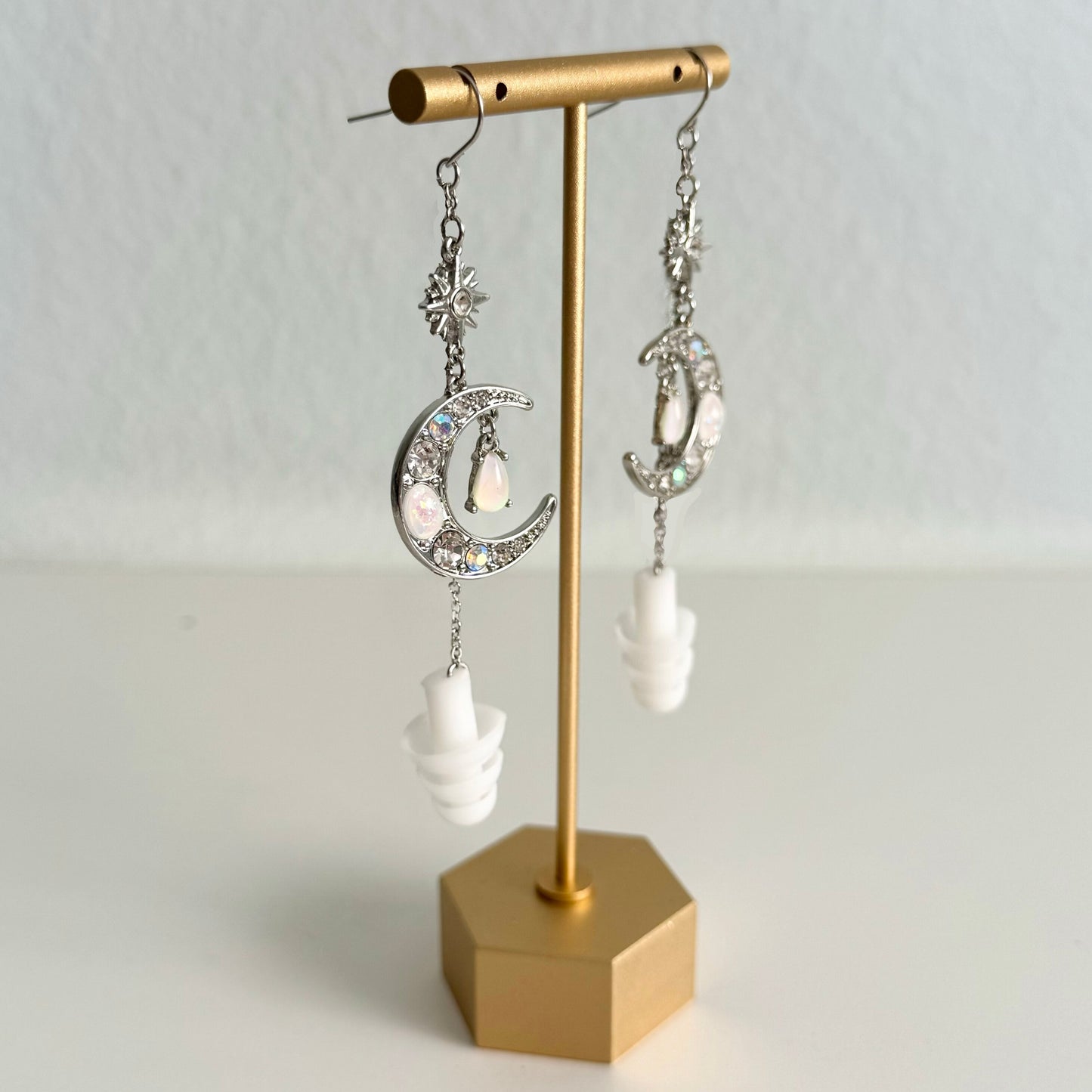 Earplug earring jewelry with crystal encrusted moons and white ear plugs attached. View of this rave accessory hanging on a gold stand.
