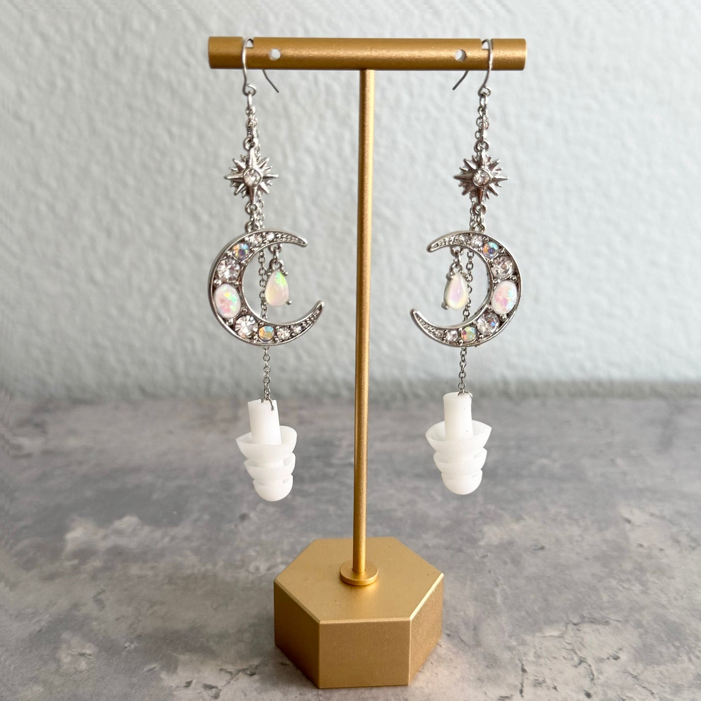 Earplug earring jewelry with crystal encrusted moons and white ear plugs attached. View of this rave accessory hanging on a gold stand.
