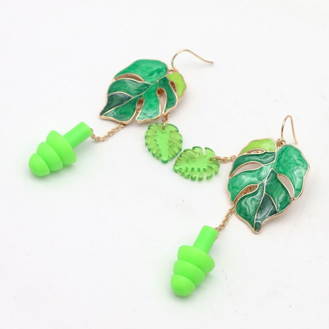 Earplug Earrings featuring Green leaf Monsteras. The earrings are laying on flat surface. The ear plugs make this jewelry great for hearing protection at raves, concerts, and other loud environments.
