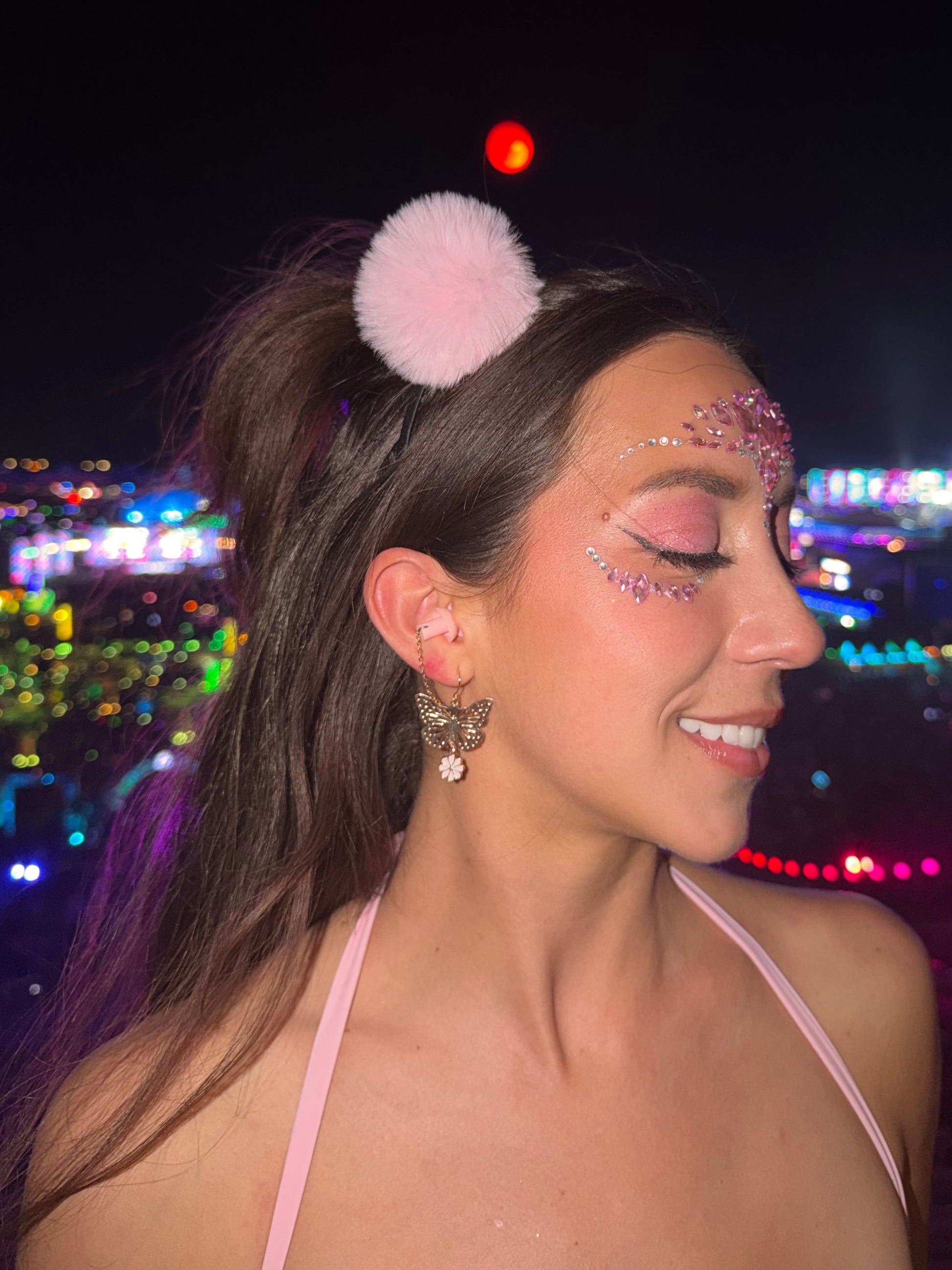 Woman with glittery makeup and pink hair accessory against a dark background with colorful lights. Modeling pink blossom earrings with ear plugs attached. 